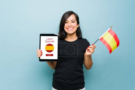 Téléchargez les photos : Belle femme caucasienne tenant le drapeau d'Espagne et montrant une tablette avec une application d'apprentissage ou un site Web pour étudier l'espagnol - en image libre de droit