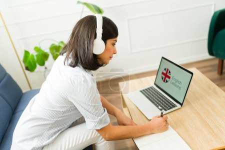 Photo for Caucasian attractive woman with headphones taking English lessons online and having a virtual class using the laptop studying - Royalty Free Image
