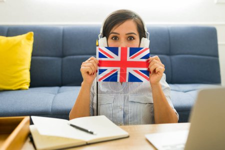 Photo for Surprised young woman with headphones excited about learning English online while holding the United Kingdom flag - Royalty Free Image