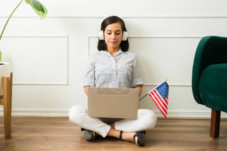 Téléchargez les photos : Belle femme caucasienne avec casque stuyding et apprendre l'anglais en ligne sur le site de l'éducation en utilisant l'ordinateur portable avec le drapeau américain - en image libre de droit