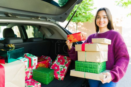 Foto de Atractiva mujer hispana sonriendo mirando a la cámara mientras lleva un montón de regalos de Navidad desde el maletero del coche después de ir de compras - Imagen libre de derechos