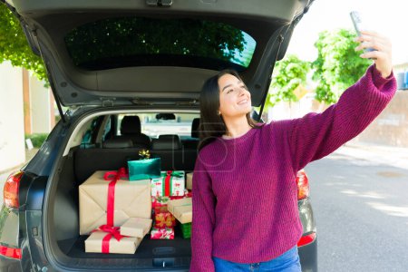 Foto de Mujer hermosa feliz usando su teléfono inteligente tomar una selfie con los regalos de Navidad en el maletero del coche después de ir de compras - Imagen libre de derechos