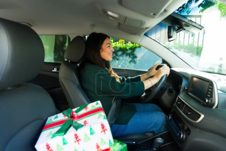 Photo for Cheerful latin woman driving and smiling with christmas presents in the car looking happy during the holidays - Royalty Free Image