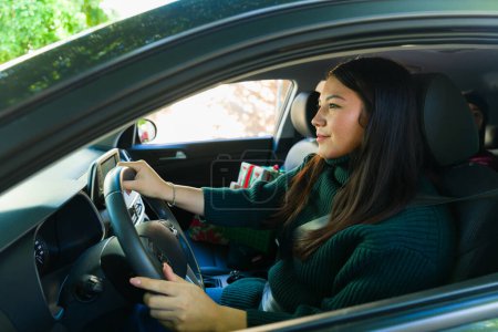 Téléchargez les photos : Latina jeune femme conduire la voiture regardant heureux tout en allant faire du shopping pour les cadeaux de Noël avec la ceinture de sécurité sur - en image libre de droit