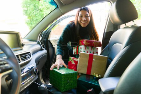 Téléchargez les photos : Heureuse femme attrayante souriant tout en conduisant et en faisant du shopping mettre les cadeaux de Noël dans la voiture pour les vacances - en image libre de droit