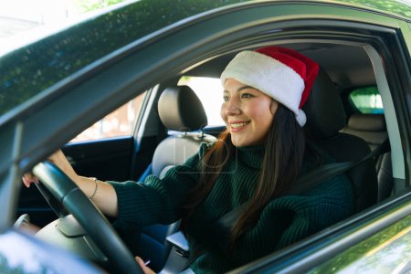 Photo for Beautiful caucasian young woman driving her car with a santa hat celebrating the holidays going shopping for christmas gifts - Royalty Free Image