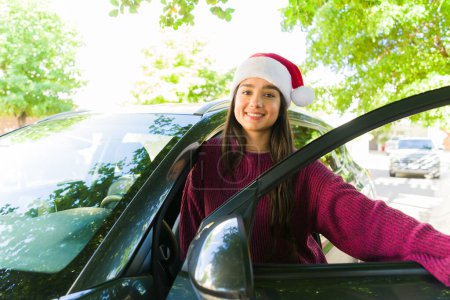 Photo for Cheerful woman in her 30s with a santa hat getting into her car to drive and shop for christmas gifts for the holiday - Royalty Free Image
