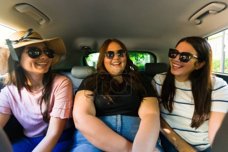 Foto de Mujeres alegres mejores amigos con gafas de sol viajando juntos en el asiento trasero del coche para un viaje de verano por carretera - Imagen libre de derechos
