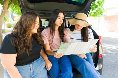 Foto de Mujeres hispanas mejores amigas sentadas en el maletero del auto mientras conducen juntas con un mapa para un divertido viaje por carretera - Imagen libre de derechos