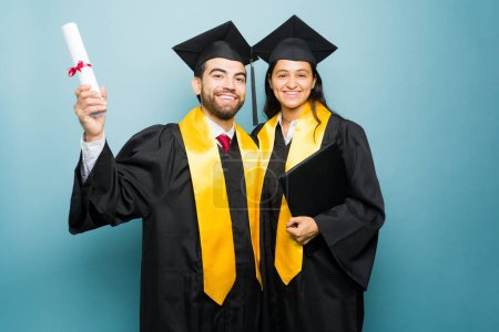 Téléchargez les photos : Heureux jeune homme excité et femme hispanique avec robe de remise des diplômes finissant le collège ensemble et montrant leur diplôme universitaire - en image libre de droit