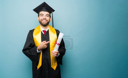 Téléchargez les photos : Attrayant heureux étudiant de l'université souriant portant sa robe de remise des diplômes et casquette après avoir reçu son diplôme d'études collégiales contre une publicité espace copie - en image libre de droit