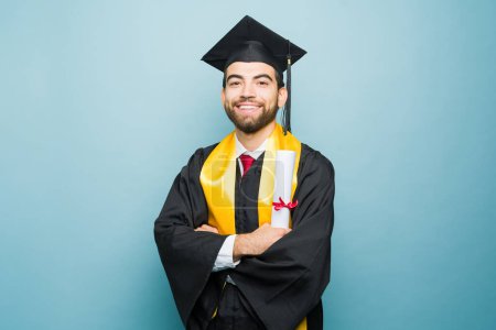 Foto de Retrato de un atractivo mexicano en su graduación universitaria sosteniendo su diploma y sonriendo después de terminar la universidad - Imagen libre de derechos