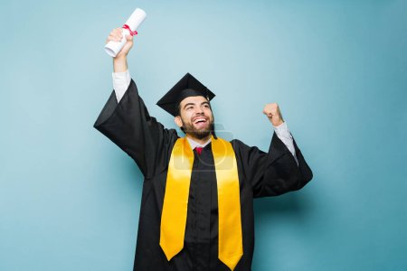 Photo for Happy excited latin man screaming and celebrating receiving his college diploma while wearing a graduation gown and cap - Royalty Free Image