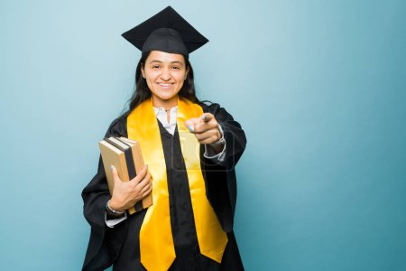 Foto de Mujer joven inteligente señalando mientras lleva libros con un vestido de graduación listo para recibir su diploma universitario después de terminar la universidad - Imagen libre de derechos