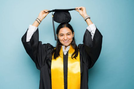 Photo for Smiling excited latin woman putting on he graduation cap and gown ready for her ceremony to receive her university diploma - Royalty Free Image