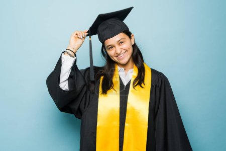 Téléchargez les photos : Mexicain heureux jeune femme mettre sa casquette de graduation et robe tout en souriant prêt à recevoir son diplôme d'études collégiales après avoir terminé l'université - en image libre de droit