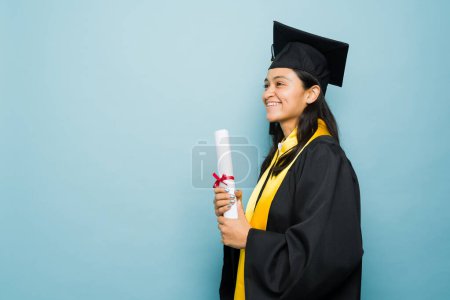 Foto de Feliz hermosa mujer latina con su vestido de graduación sonriendo después de recibir su diploma universitario en un fondo con espacio de copia - Imagen libre de derechos