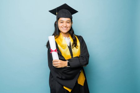 Foto de Joven mexicana sonriendo feliz por terminar la universidad usando su vestido de graduación para recibir su diploma universitario - Imagen libre de derechos
