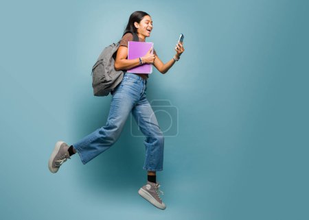 Téléchargez les photos : Toute la longueur d'une belle femme latine sautant dans les airs en s'amusant tout en allant à l'université portant son sac à dos sur un fond bleu studio - en image libre de droit