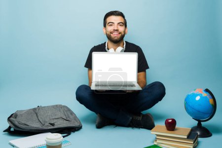 Téléchargez les photos : Heureux étudiant mexicain homme montrant l'écran d'ordinateur portable vierge et souriant tout en étudiant pour l'université en ligne avec beaucoup de livres - en image libre de droit