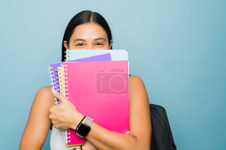Photo for Hispanic young woman student hiding behind her college notebooks looking happy studying university - Royalty Free Image
