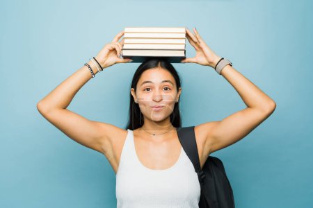Foto de Mujer joven latina atractiva inteligente sosteniendo muchos libros sobre su cabeza mientras estudia para la universidad haciendo tarea - Imagen libre de derechos