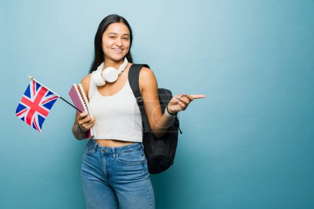 Téléchargez les photos : Femme hispanique joyeuse avec le drapeau du Royaume-Uni apprendre l'anglais à l'université pour étudier à l'étranger tout en pointant vers copier l'espace sur un fond bleu - en image libre de droit