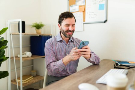 Photo for Latin happy entrepreneur or lawyer smiling while texting on the smartphone while working at his office desk - Royalty Free Image