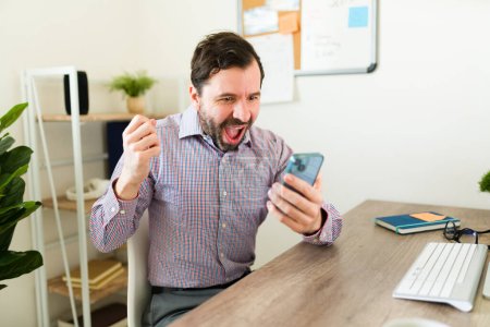 Photo for Cheerful happy entrepreneur and boss receiving good news while working at the office and using the smartphone celebrating - Royalty Free Image