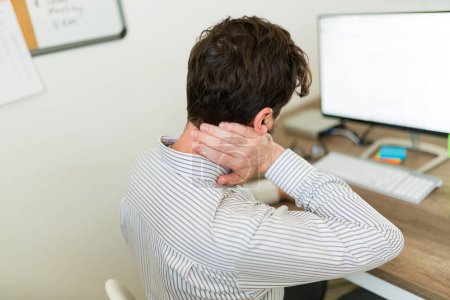 Photo for Hispanic lawyer and boss rubbing his neck feeling tired and stressed seen from behind working at his desk - Royalty Free Image