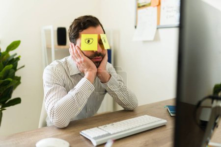 Téléchargez les photos : Homme d'affaires endormi dormant assis à son bureau en mettant des notes collantes sur ses yeux et en travaillant - en image libre de droit