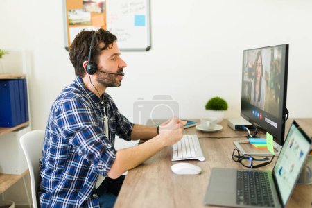 Photo for Side view of a sales representative with a headset sitting at the office desk and having a work video call - Royalty Free Image