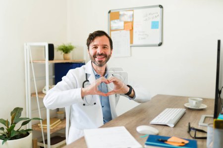Photo for Cheerful cardiologist doctor making a heart love gesture and smiling working at his office waiting for a medical examination - Royalty Free Image