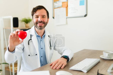 Photo for Handsome latin cardiologist doctor showing a healthy heart and smiling with a lab coat and stethoscope at his office - Royalty Free Image