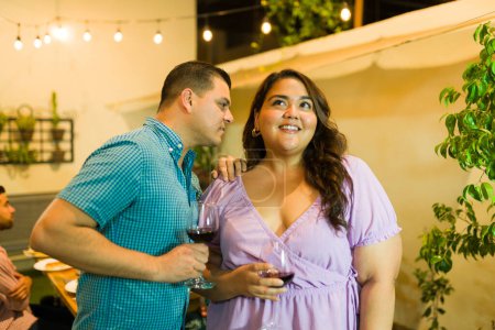 Foto de Hombre hispano susurrando un secreto a una joven bebiendo vino y coqueteando durante una celebración nocturna con amigos - Imagen libre de derechos