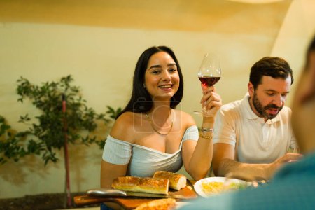 Photo for Attractive young woman smiling making eye contact while drinking wine and eating dinner with her group of friends - Royalty Free Image