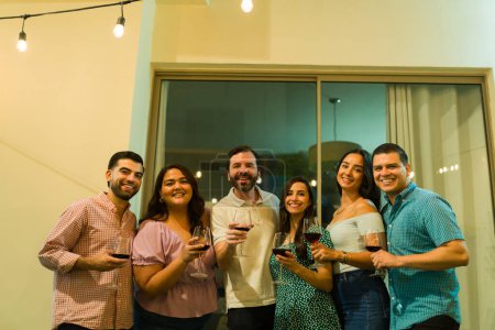 Foto de Grupo hispano de personas bebiendo vino después de cenar celebrando con amigos en el patio trasero por la noche - Imagen libre de derechos