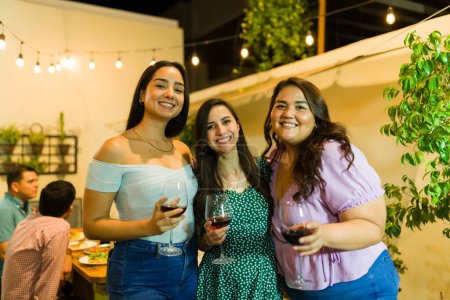 Téléchargez les photos : De belles jeunes femmes souriantes buvant du vin lors d'un dîner avec des amis dans la cour pendant la nuit - en image libre de droit