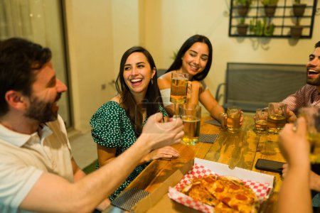 Foto de Hermosas mujeres y hombres riendo diciendo vítores y haciendo un brindis con amigos mientras se divierten bebiendo cerveza y comiendo pizza - Imagen libre de derechos