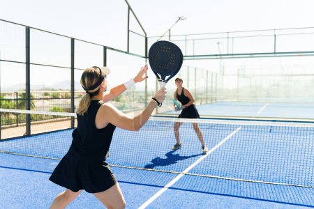 Foto de Pareja de mujeres caucásicas de mediana edad jugando un partido de padel mientras hacen ejercicio juntas en la cancha de tenis - Imagen libre de derechos