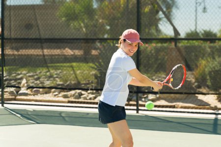 Téléchargez les photos : Jeune femme active excitée souriant et s'amusant tout en jouant à un jeu de tennis avec des amis pendant une séance d'entraînement et en faisant un revers - en image libre de droit