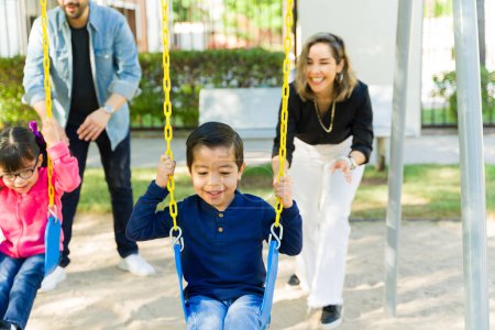 Photo for Cheerful parents playing with their kids in the park and pushing them on the swings while having fun together - Royalty Free Image