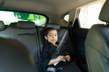 Téléchargez les photos : Adorable petit garçon caucasien souriant faisant un contact visuel tout en étant assis sur le siège arrière de la voiture à l'aide d'un siège d'appoint va à la classe de karaté - en image libre de droit