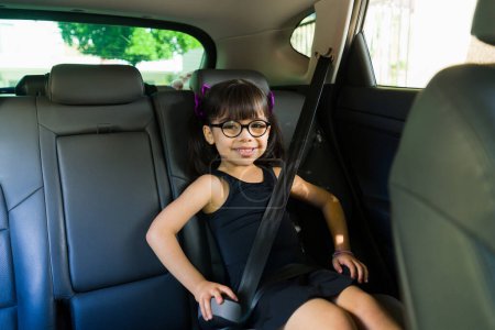Téléchargez les photos : Belle petite fille caucasienne avec des lunettes souriant assis dans le siège d'appoint de la voiture tout en allant à la classe de danse de ballet - en image libre de droit