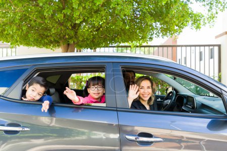 Photo for Excited parents and family of four with children smiling waving hello while driving in the car having fun - Royalty Free Image