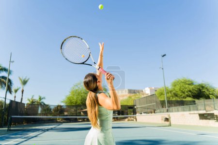 Foto de Hermosa mujer latina vista desde atrás lanzando una pelota de tenis en el aire para practicar su servicio mientras juega al tenis - Imagen libre de derechos