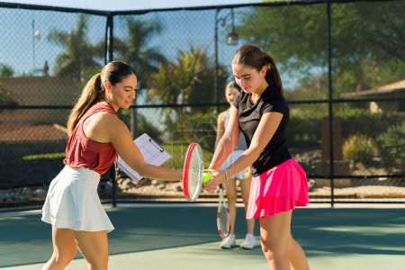 Foto de Feliz entrenador latino dando lecciones a las adolescentes que enseñan a jugar al tenis y utilizar raquetas en la cancha de tenis - Imagen libre de derechos