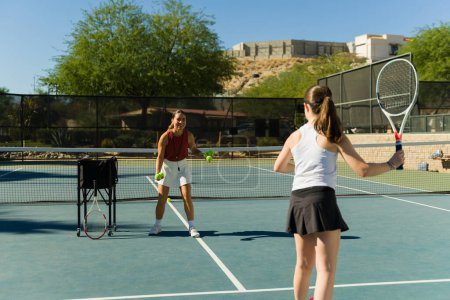 Foto de Mujer latina alegre y entrenador de tenis lanzando una pelota a una adolescente activa tomando clases de tenis - Imagen libre de derechos