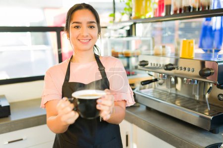Foto de Retrato de una atractiva mujer latina con un delantal trabajando como camarera entregando café a un cliente y sonriendo en la cafetería - Imagen libre de derechos