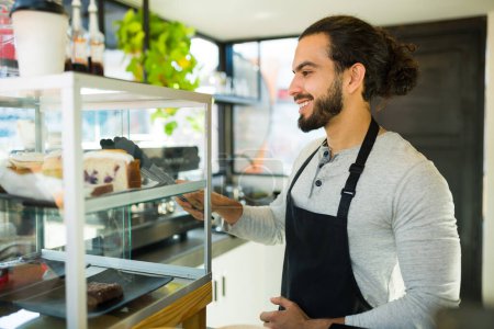 Photo for Attractive hispanic young man smiling working as a barista handing sweet dessert to a customer at the coffee shop - Royalty Free Image
